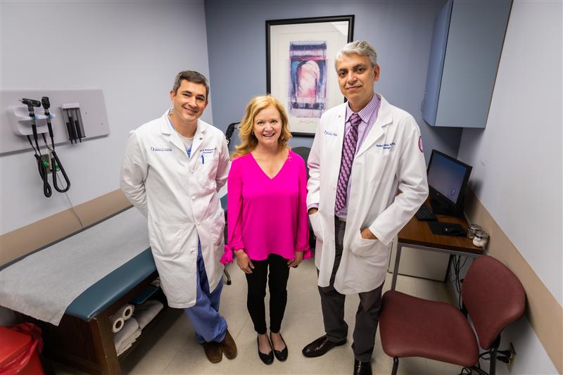 A smiling stroke patient standing between two doctors in an exam room.